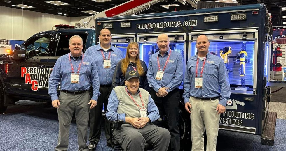 A group of six people, five standing and one seated, pose in front of a truck with PAC branding. They wear matching blue shirts and conference badges, smiling warmly.
